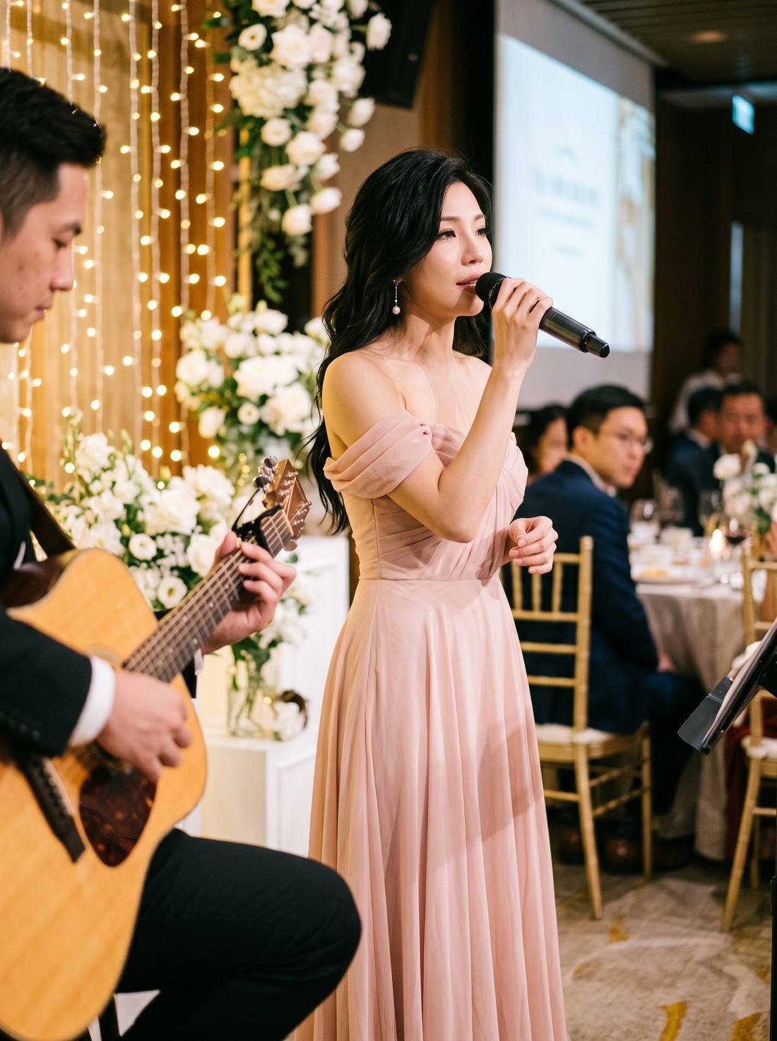 Pauline Tan in a blush chiffon gown singing at a wedding dinner beside white flowers, with a guitarist seated nearby.